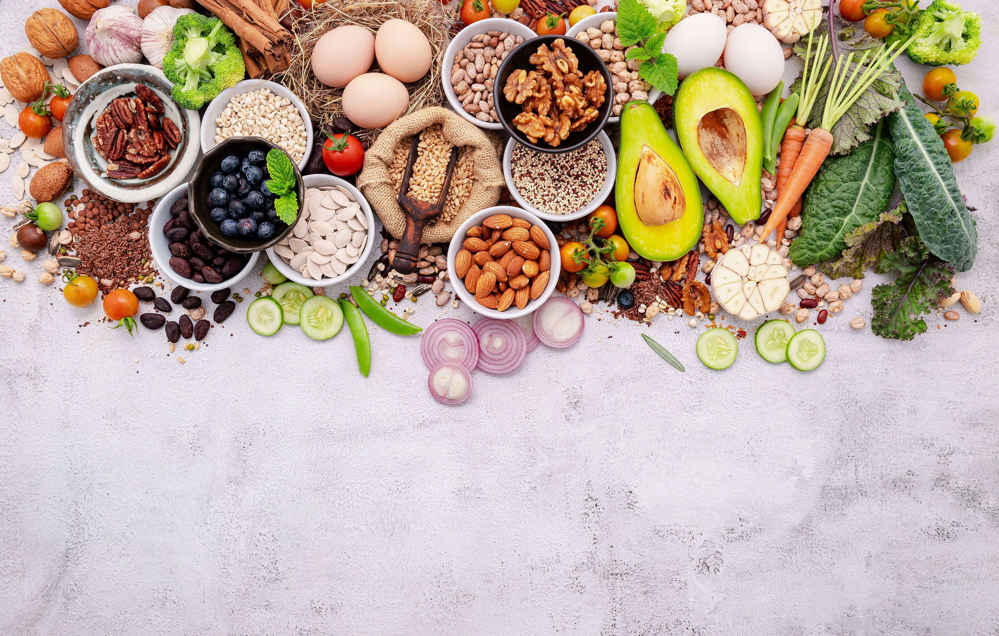 ACGrace - Assorted healthy foods including nuts, seeds, eggs, avocado, vegetables, leafy greens, and berries arranged on a light surface with empty space below.