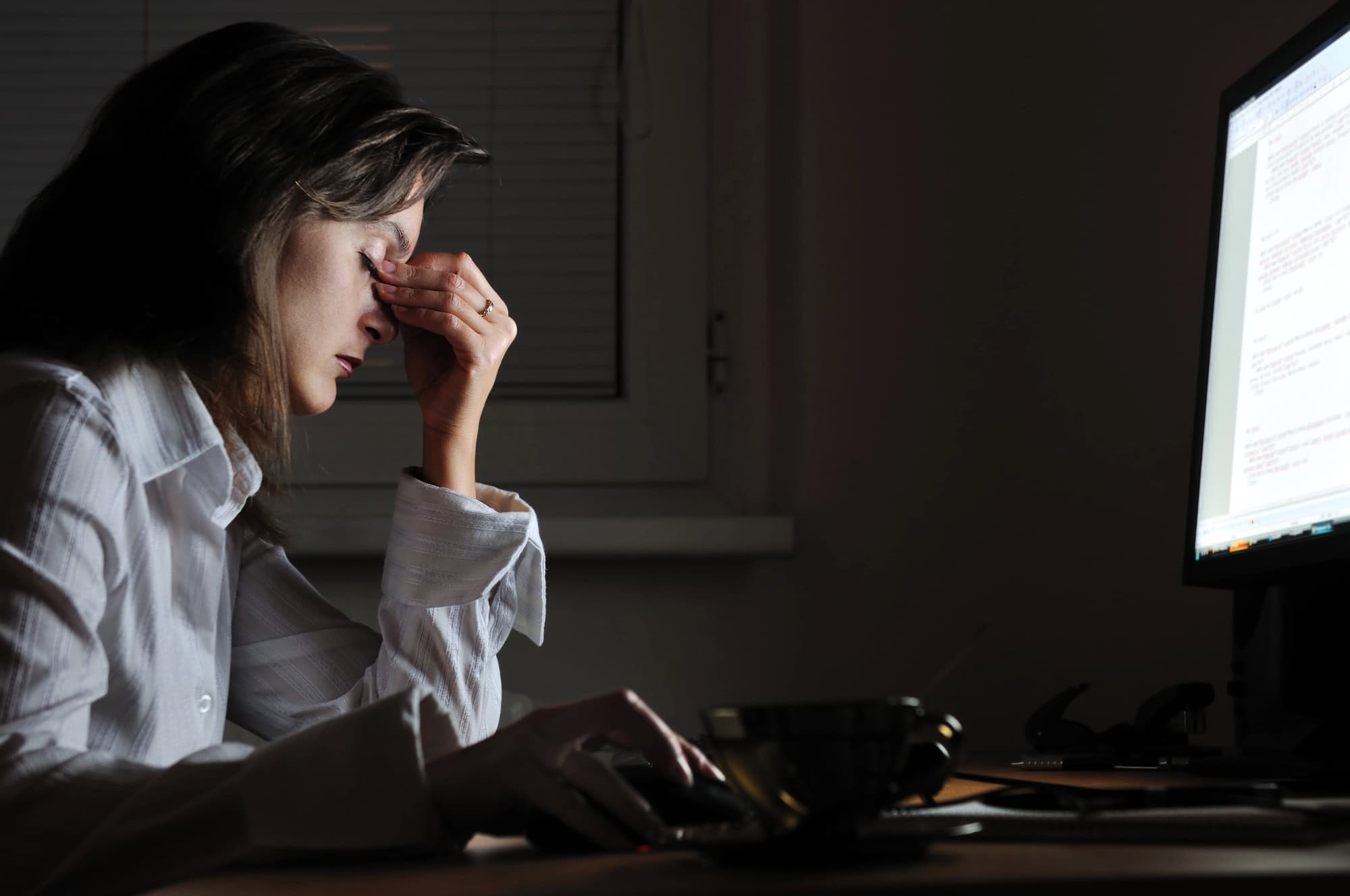ACGrace - A person sits at a desk in front of a computer screen, rubbing their eyes with a tired expression, suggesting fatigue or stress while working late.