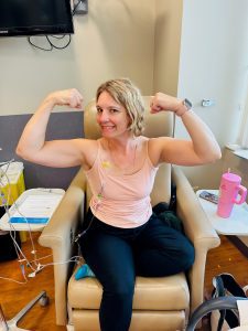 ACGrace - A woman sits in a medical chair with IV lines attached, flexing her arms and smiling. Medical supplies and a pink cup are on a table beside her.