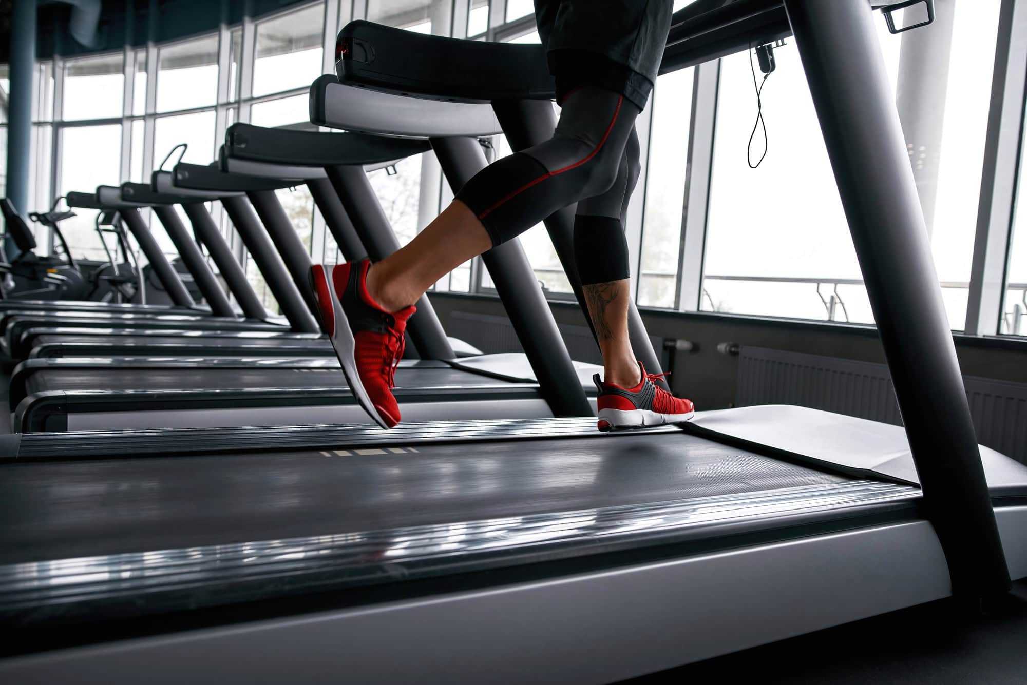 ACGrace - Person running on a treadmill in a gym, wearing black leggings and red sneakers, with several other treadmills lined up nearby.