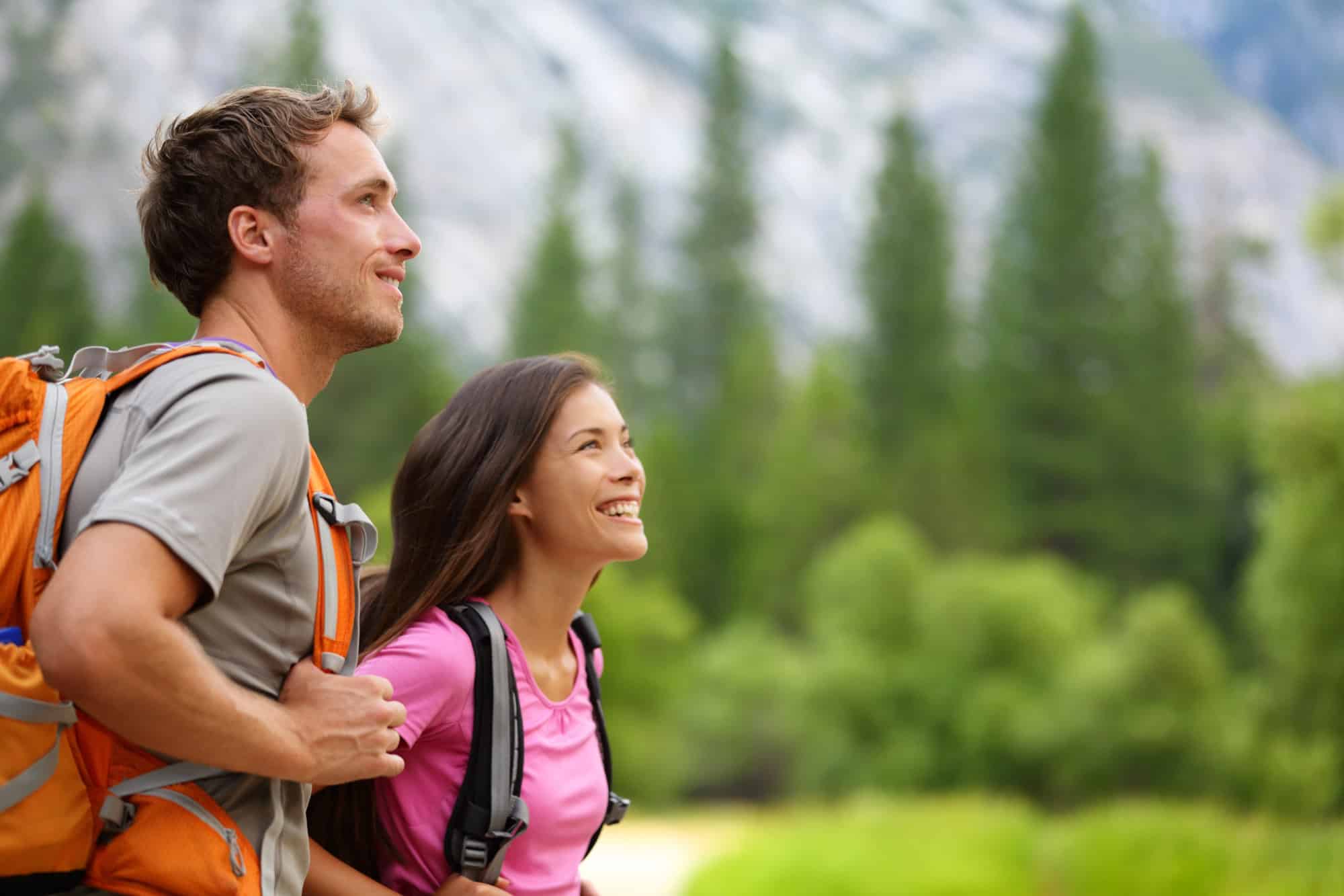 ACGrace - A man and woman wearing backpacks are hiking outdoors, with trees and mountains in the blurred background.