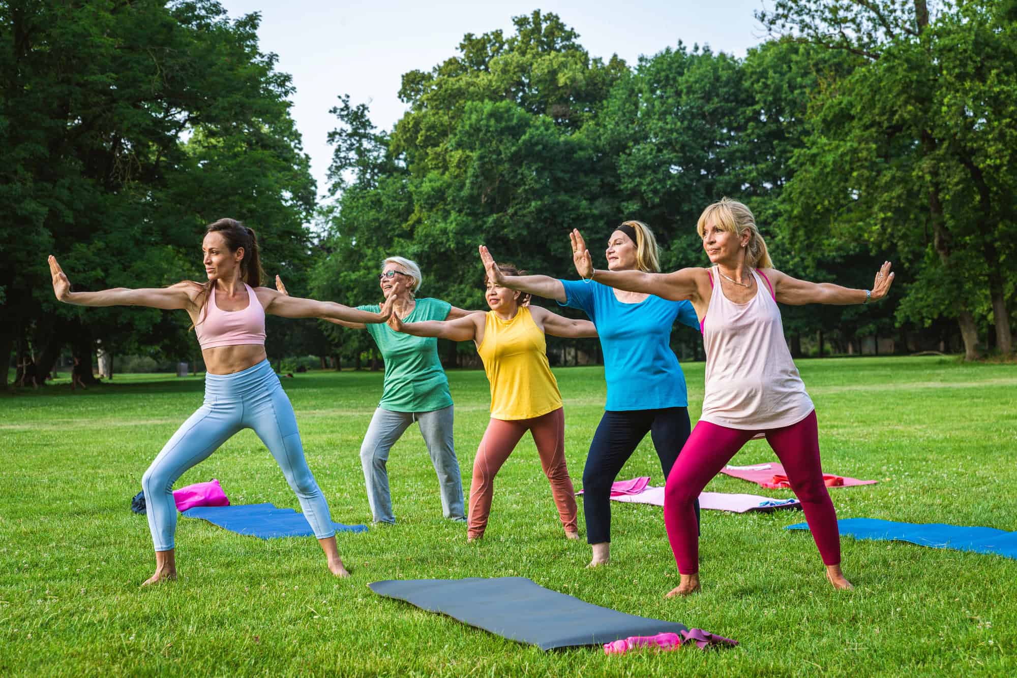 ACGrace - Five women practicing yoga together outdoors on a grassy field, each holding a warrior pose with arms extended, yoga mats on the ground, surrounded by trees.