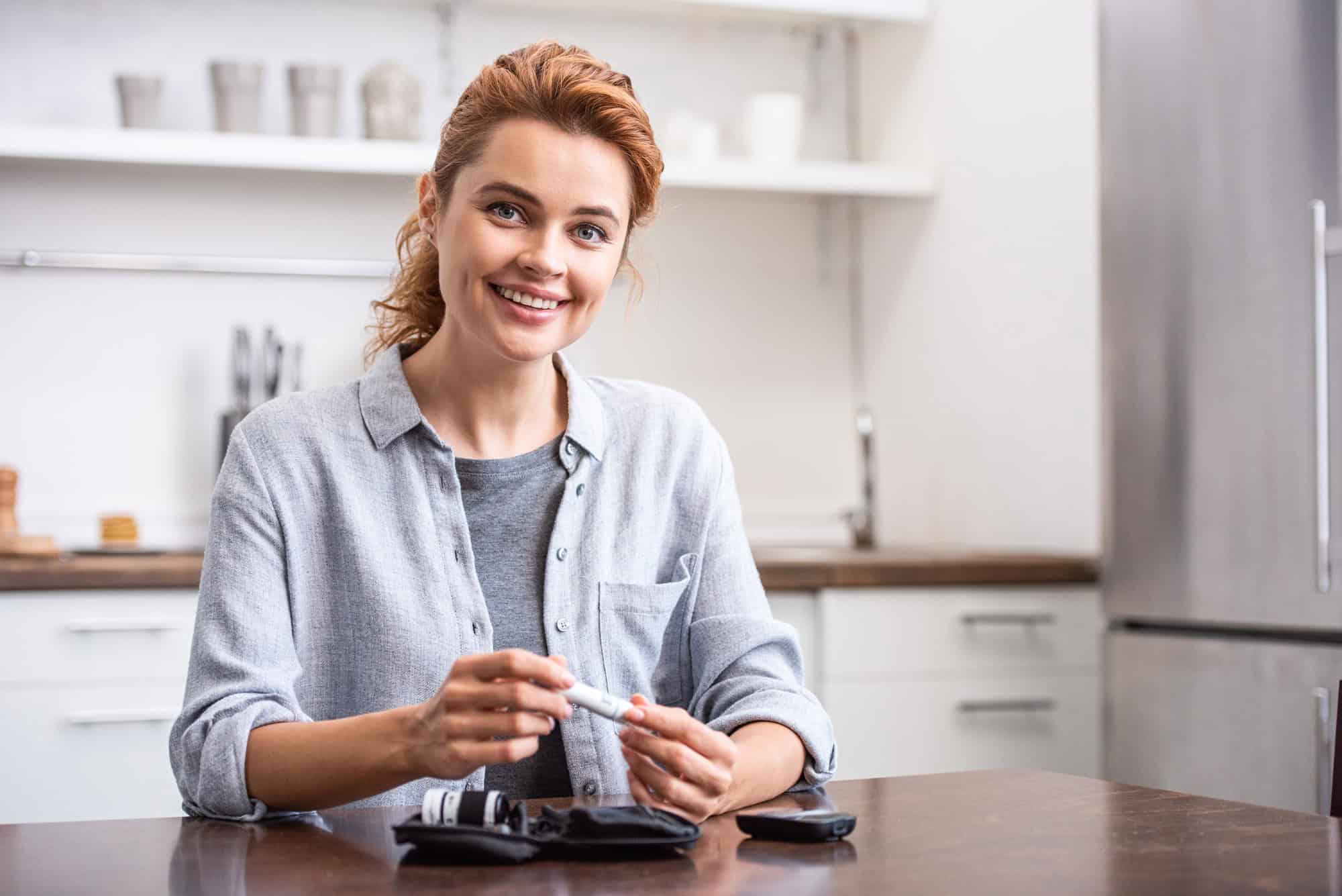 ACGrace - Smiling woman sits at a kitchen table preparing a blood glucose meter with supplies laid out in front of her.